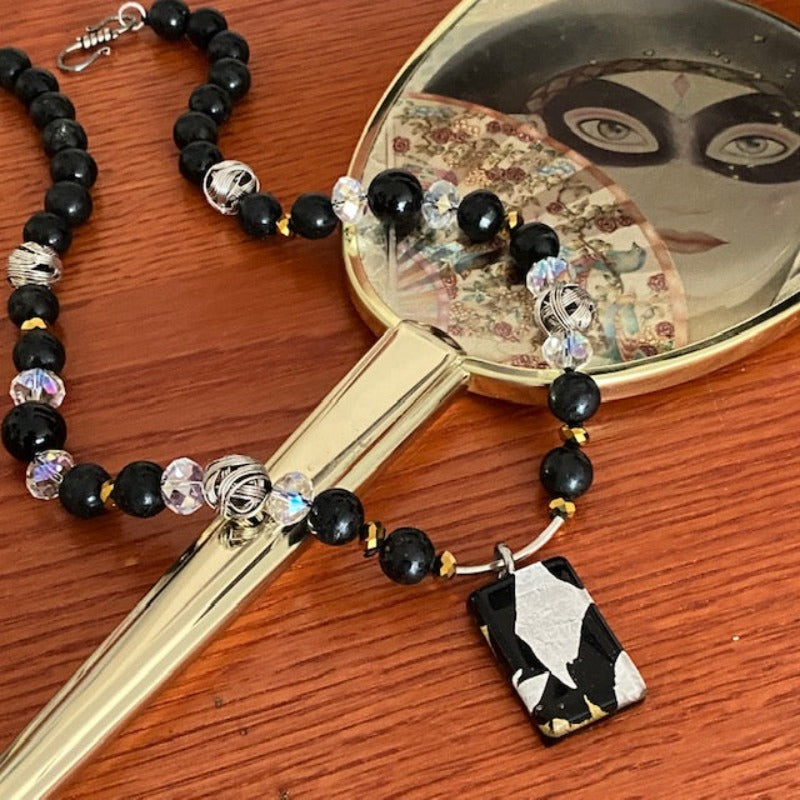 Black-silver-gold Dichroic Glass 18-inch kinetic necklace shown draped on an old-fashioned mirror with stylized masked woman's face